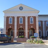 Front view of the Delaware Court of Chancery in Georgetown, Delaware, on a sunny day. The building features a red brick facade with white columns, arched entryways, and the state seal above the inscription “Court of Chancery”; a historical marker and parked vehicles are visible in the foreground.