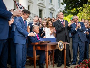 A press photo of President Donald Trump sitting at a desk holding a signed document, surrounded by applauding people in formal attire outside the White House.